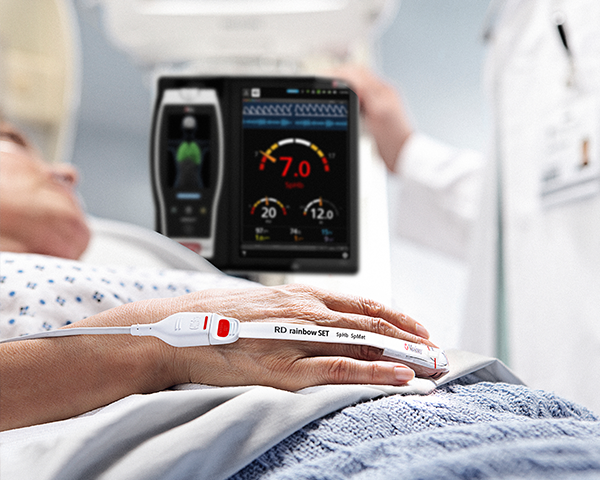 Paziente in un letto d'ospedale monitorato tramite sensore Masimo con dispositivo Root e medico sullo sfondo che consulta il dispositivo Root.