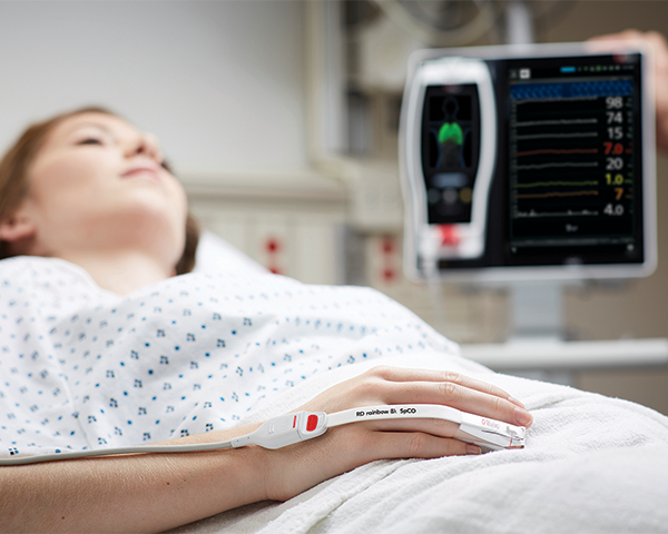 Paziente addormentato in un letto d'ospedale che indossa un sensore da dito Masimo con dispositivo Root sullo sfondo.